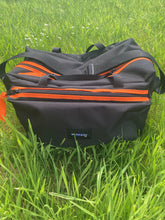 Load image into Gallery viewer, Small black and mad orange Midwater Carryall with carry handles and a shoulder strap, showing external pocket, placed on a grassy field.